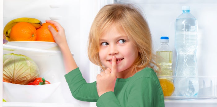 Niña rubia haciendo la señal de silencio mientras saca snacks saludables de la nevera