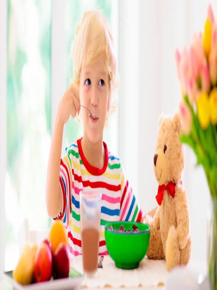 Niño rubio desayunando junto a algunas frutas y un oso de peluche